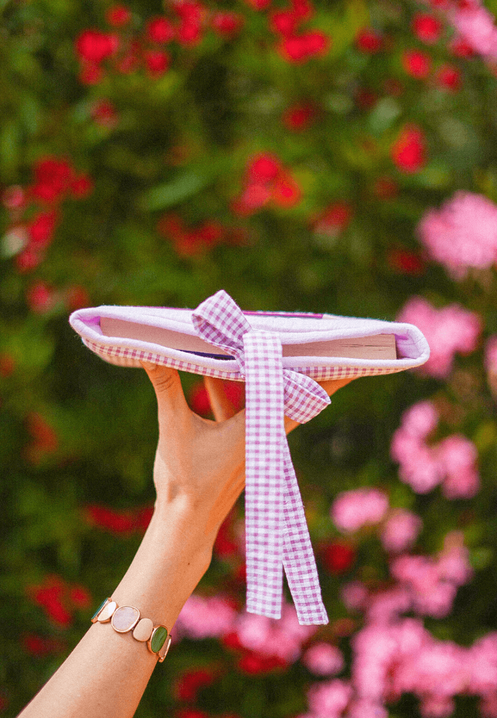 Funda personalizada para libro de bolsillo Lavander Vichy, artesanal y acolchadita, sujetada con una mano frente a flores.